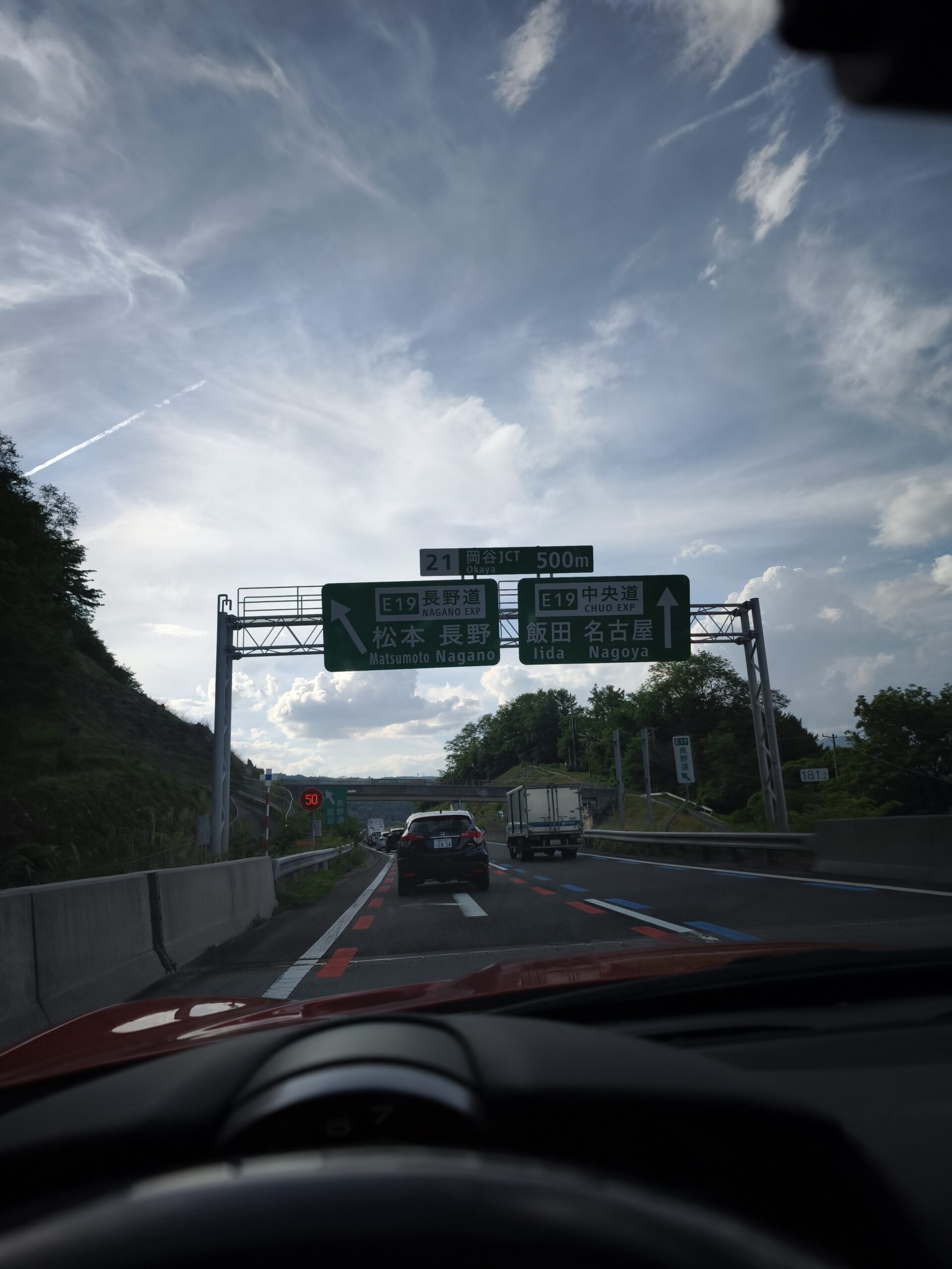 MVIMG_20260619_165006 – mbFAST Tuning Highway scene with large green overhead signs indicating exits toward Matsumoto, Nagano, Iida, and Nagoya; cars on the road beneath a blue sky with clouds.