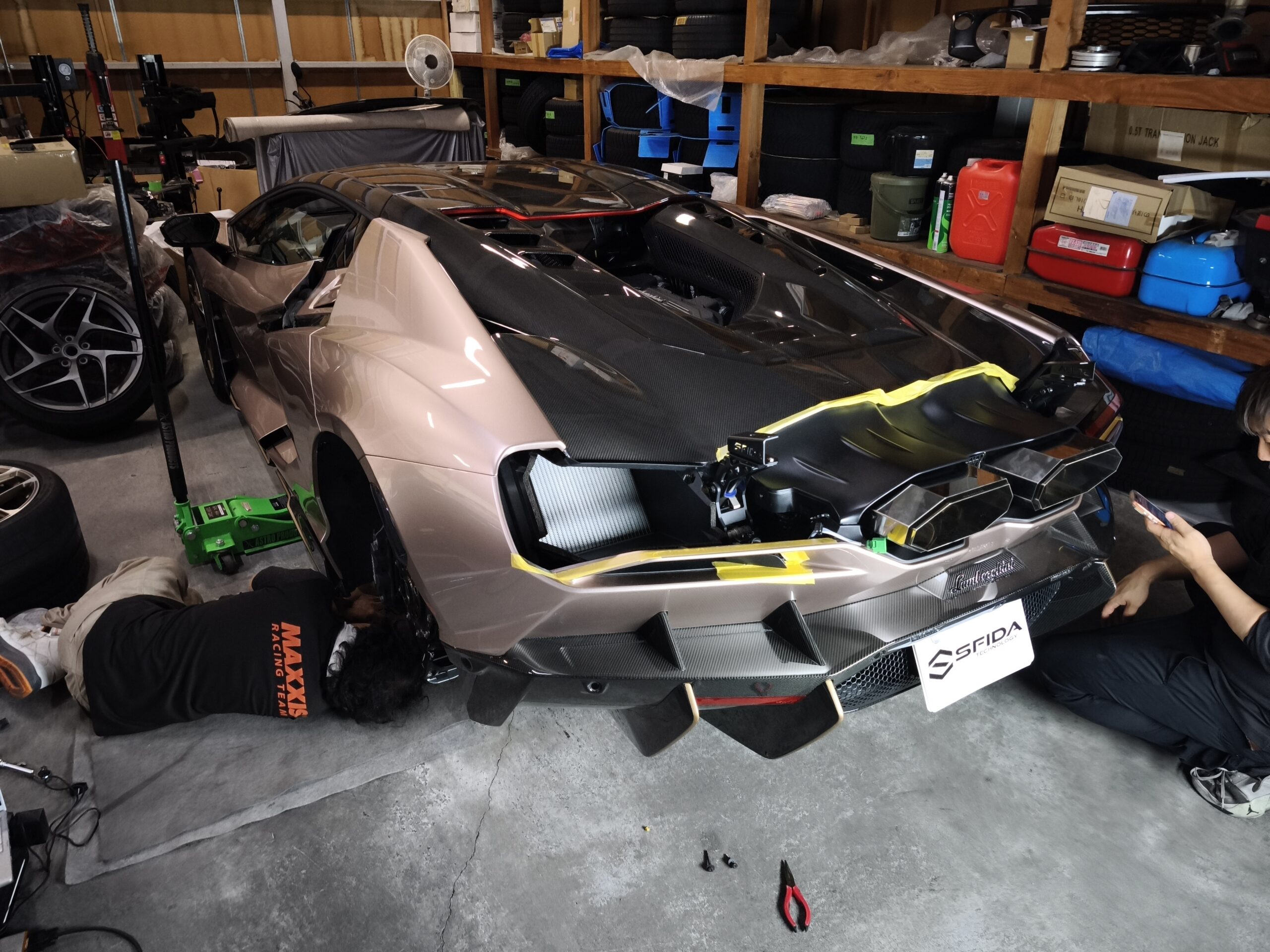MVIMG_20260619_202543 – mbFAST Tuning Silver Lamborghini Aventador being serviced in a cluttered home garage; technicians work near the front with tools on the floor and shelves full of tires and boxes nearby.
