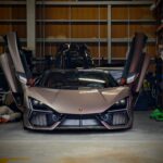 Front view of a silver Lamborghini with upward-opening doors in a cluttered garage/workshop setting