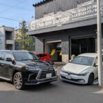 Garage front with sign 'Charism Garage' and two parked cars (black Lexus SUV and white sedan) on a sunny street.