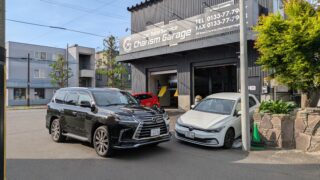 PXL_20260603_073251846 – mbFAST Tuning Garage front with sign 'Charism Garage' and two parked cars (black Lexus SUV and white sedan) on a sunny street.