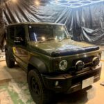 Three-quarter front view of a dark green Jeep Wrangler with off-road lights, parked inside a studio-like space with black plastic walls and hexagonal neon ceiling lights.