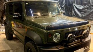 WhatsApp Image 2026-05-27 at 211800 – mbFAST Tuning Three-quarter front view of a dark green Jeep Wrangler with off-road lights, parked inside a studio-like space with black plastic walls and hexagonal neon ceiling lights.