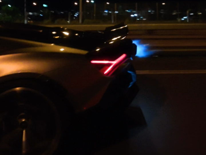 image – mbFAST Tuning Nighttime rear view of a dark sports car with a distinctive red V-shaped taillight and blue exhaust glow on a highway edge.
