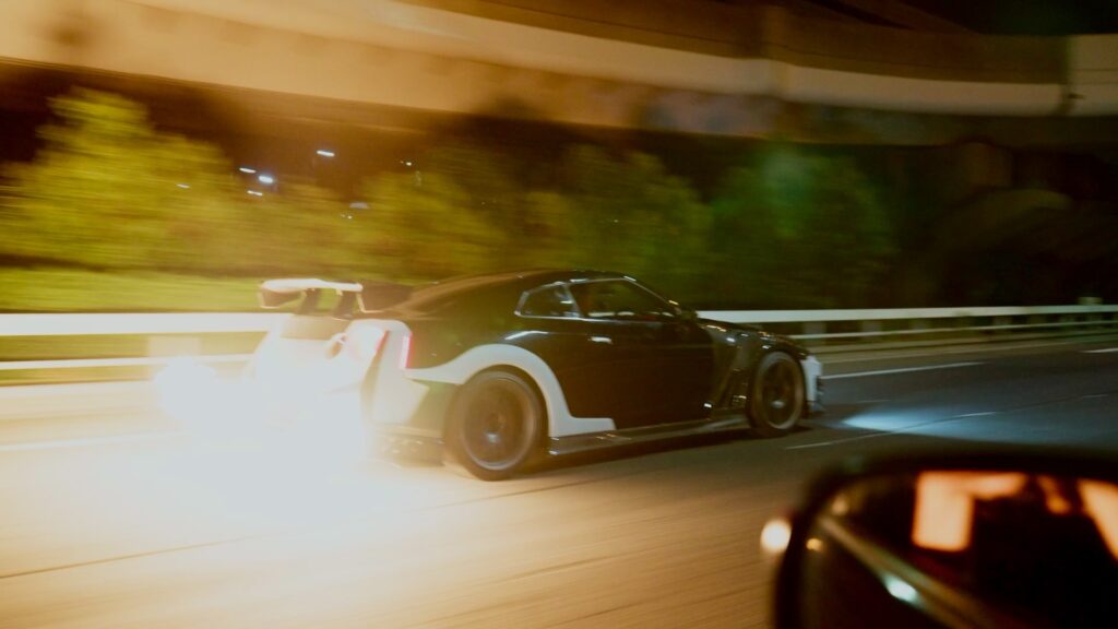 画像 24 – mbFAST Tuning Nighttime highway scene with a black sports car speeding by in motion blur, guardrail and greenery in the background.