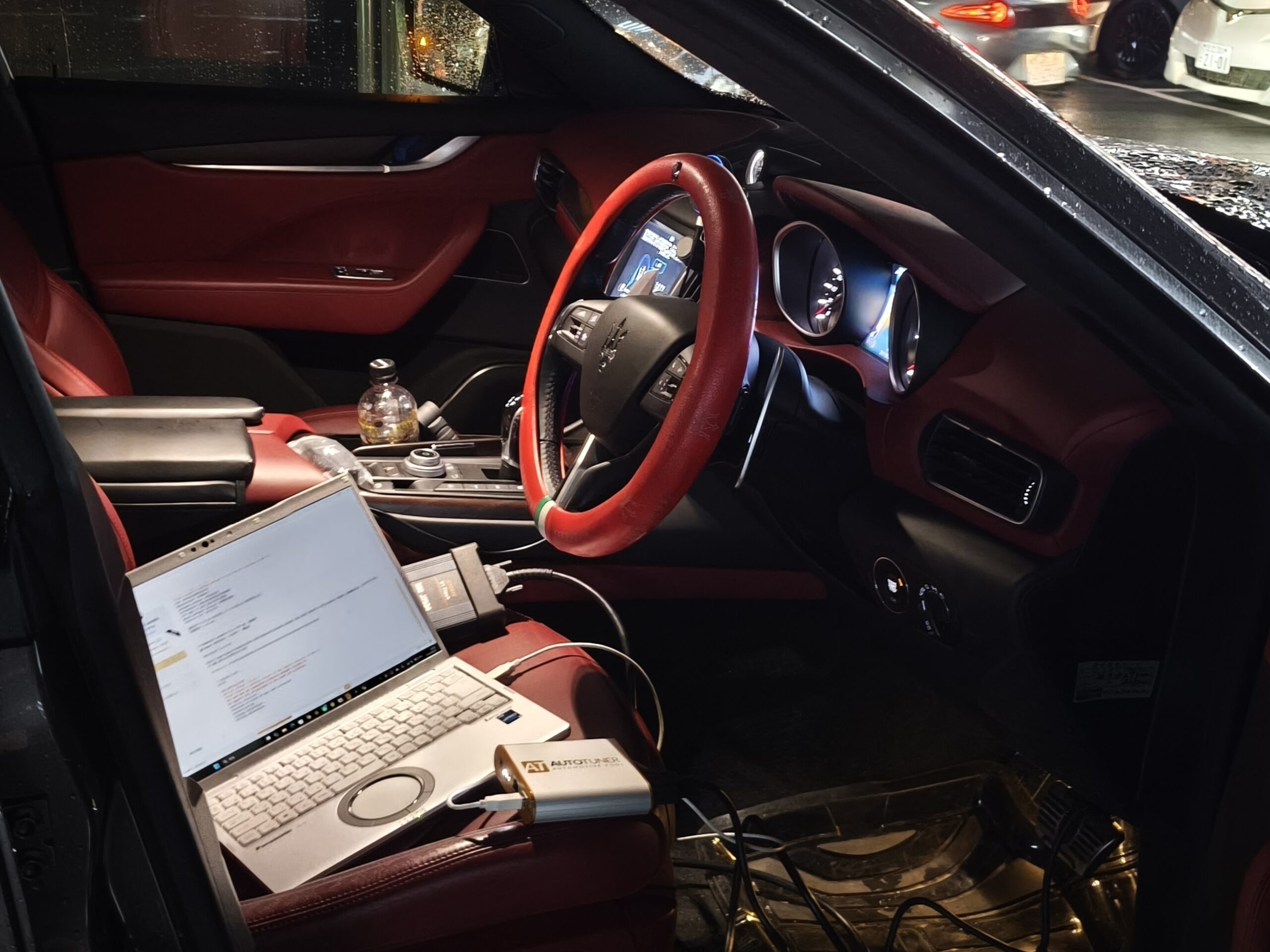 IMG_20260627_163430 – mbFAST Tuning Inside a red-leather luxury car: dashboard screens, steering wheel with red cover, laptop on the passenger seat, cables and a water bottle nearby.