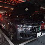 Black Maserati Levante SUV with its hood raised, parked on a metal ramp in a dim indoor parking structure with other vehicles nearby.