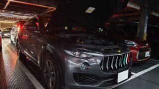 Black Maserati Levante SUV with its hood raised, parked on a metal ramp in a dim indoor parking structure with other vehicles nearby.