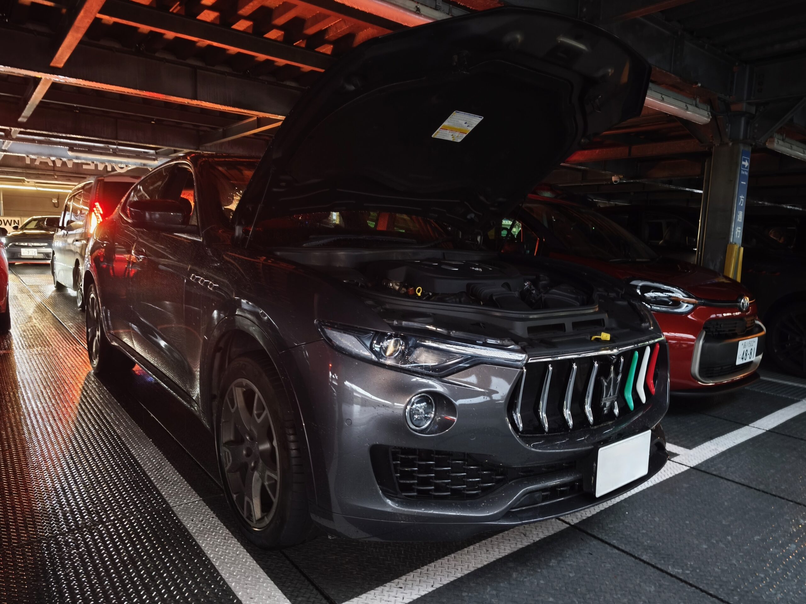 Black Maserati Levante SUV with its hood raised, parked on a metal ramp in a dim indoor parking structure with other vehicles nearby.