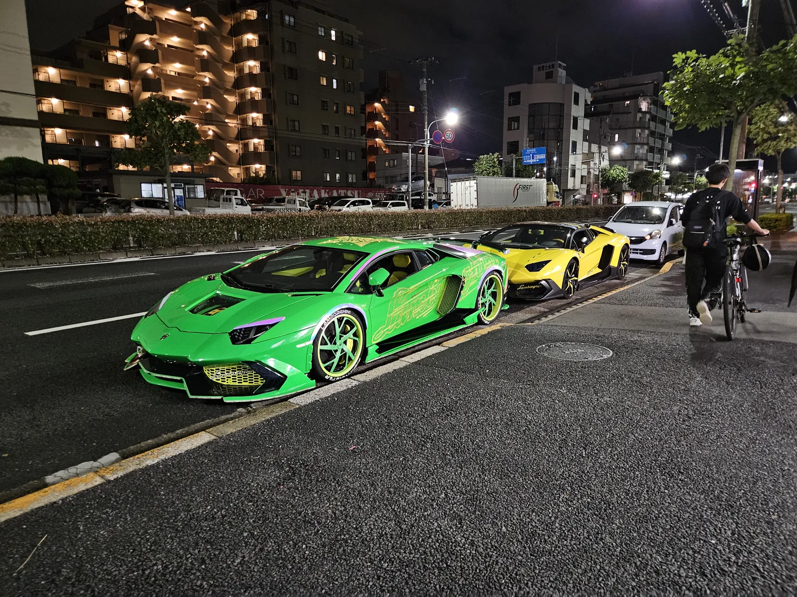 Night street at night with bright green Lamborghini in the foreground and a yellow Lamborghini behind it, a cyclist on the right, and lit apartment buildings in the background.