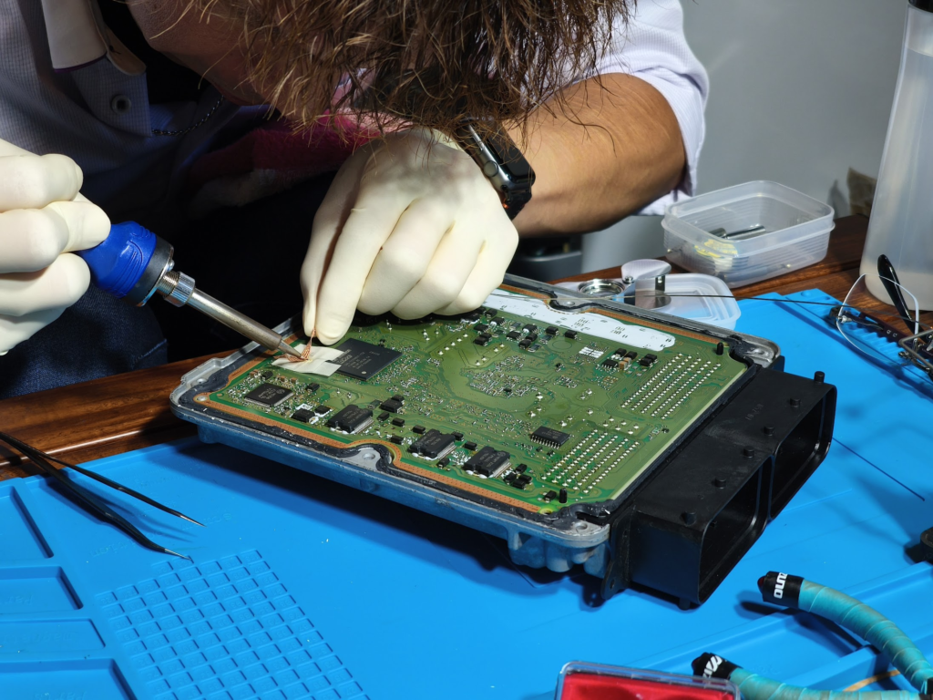 image – mbFAST Tuning Technician wearing gloves soldering a circuit board with a soldering iron on a blue work mat.