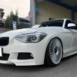 White BMW with lowered suspension and oversized multi-spoke wheels parked outside a shop or dealership bay, front angle view