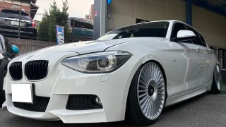 116i-f20-ecu-tuning-1 – mbFAST Tuning White BMW with lowered suspension and oversized multi-spoke wheels parked outside a shop or dealership bay, front angle view