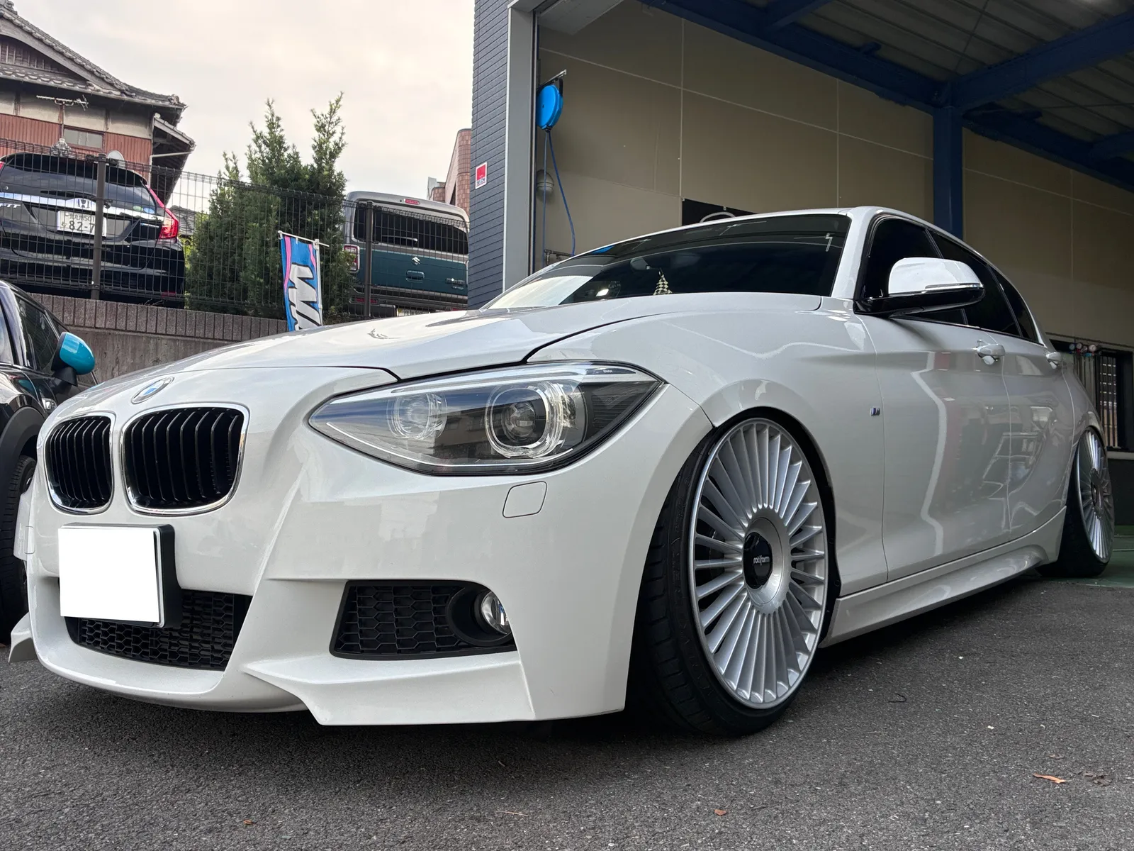 White BMW with lowered suspension and oversized multi-spoke wheels parked outside a shop or dealership bay, front angle view