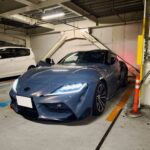 Blue sports car parked in an indoor parking garage with a white van visible in the background and yellow safety posts nearby.