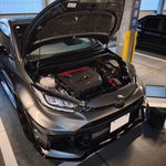 Grey SUV with its hood open, exposing the engine, in a vehicle inspection bay; a laptop sits on a stand nearby as a technician works.