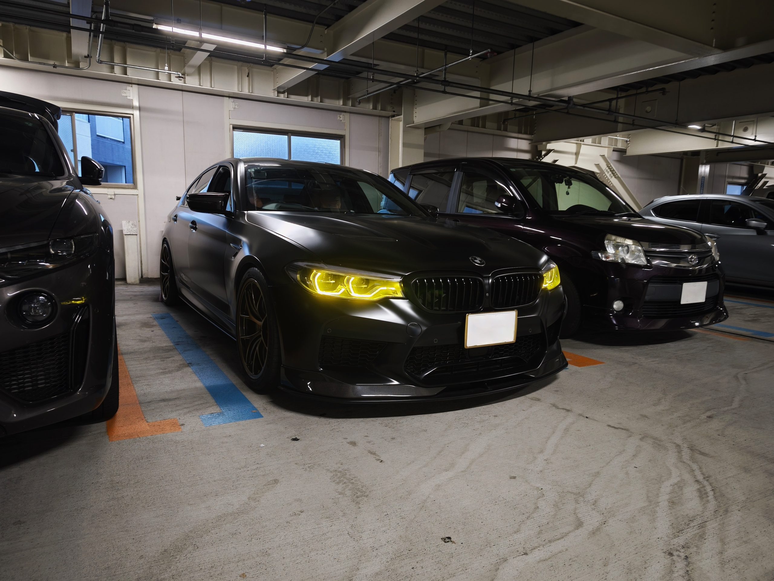 MVIMG_20260814_181851 – mbFAST Tuning Indoor parking garage with several parked cars, including a dark BMW in the foreground and a row of vehicles in the background.