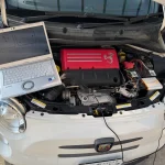 Open hood of a white Abarth car with a laptop resting on the engine bay, revealing a red engine cover and exposed wiring under the hood.