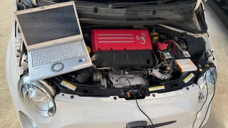 Open hood of a white Abarth car with a laptop resting on the engine bay, revealing a red engine cover and exposed wiring under the hood.