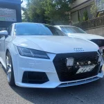 White Audi sports car front view with honeycomb grille and aftermarket LED light panels, Japanese license plate visible in a parking lot.