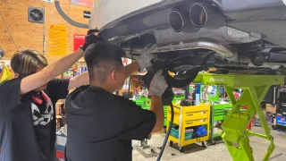 f30-oneoff-muffler-1 – mbFAST Tuning リフトアップされた車両下部でスタッフ2名がワンオフマフラーの取り付け作業を行っている様子