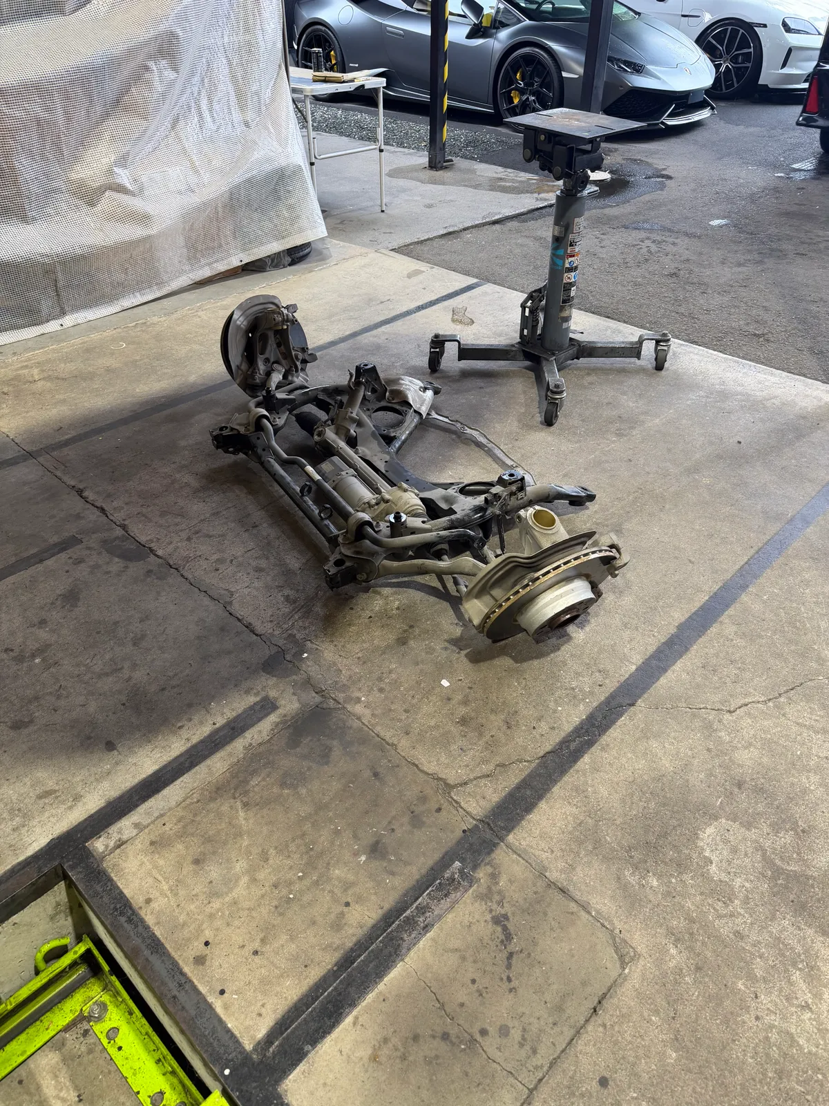f31-328i-oil-pan-replacement-3 – mbFAST Tuning Disassembled car suspension and brake rotor laid out on a concrete shop floor beside a rolling jack stand, with luxury cars visible in the background.