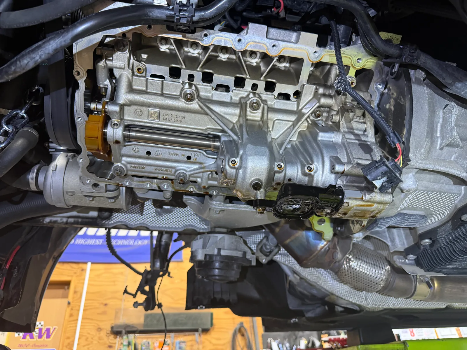 f31-328i-oil-pan-replacement-5 – mbFAST Tuning Under-vehicle view of a car engine with the valve cover removed, exposing the valve train and camshaft area in a workshop setting.
