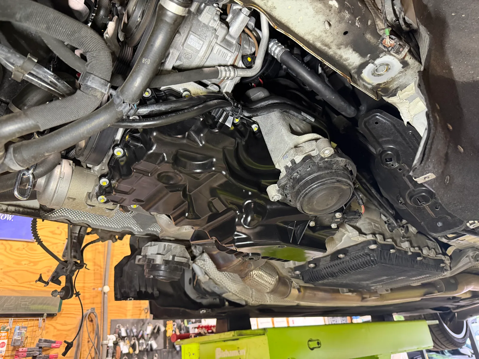 f31-328i-oil-pan-replacement-7 – mbFAST Tuning Underside of a car showing engine, belts, hoses, and suspension components in a workshop.