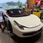 A man lying on the workshop floor under a white Ferrari, working on its undercarriage in a busy garage.