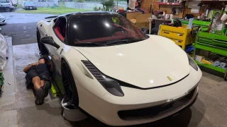ferrari458-maintenance-1 – mbFAST Tuning A man lying on the workshop floor under a white Ferrari, working on its undercarriage in a busy garage.