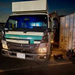 Front view of a white delivery truck with teal stripe, parked in a lot beside a large white container.