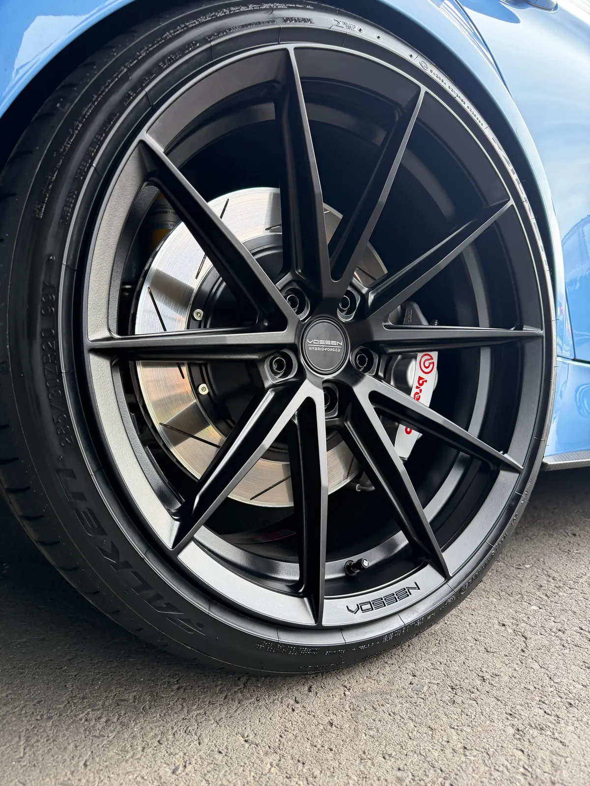 Close-up of a blue car's wheel: black multi-spoke Vossen alloy rim with a visible brake rotor and caliper behind it