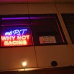 Storefront window at night with neon sign saying 'mb PIT WHY NOT RACING' and an 'We are open' sign in the window illuminated by purple and red lights.