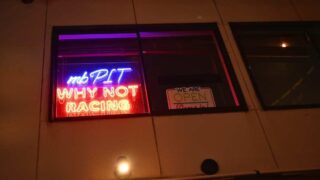 mbpit-neon-sign-shibuya – mbFAST Tuning Storefront window at night with neon sign saying 'mb PIT WHY NOT RACING' and an 'We are open' sign in the window illuminated by purple and red lights.