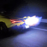 Yellow sports car on a dark highway with its rear end damaged and bright flames spewing from the exhaust at night by a guardrail