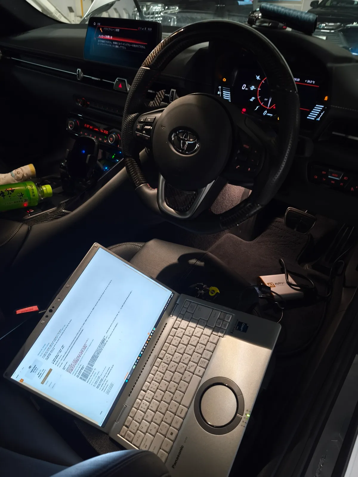 Person working in a car: laptop open on the passenger seat with dashboard lights on and steering wheel in view (car parked).