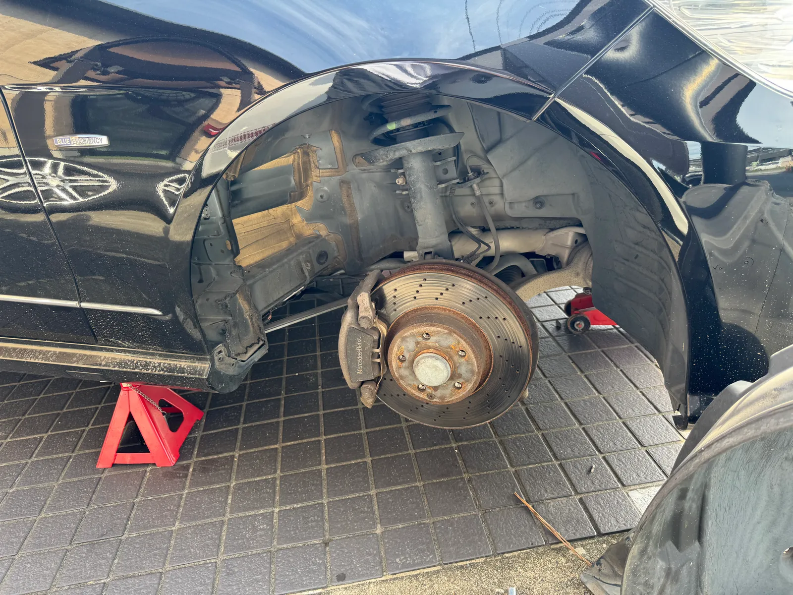 w212-e350bluetec-ecu-tuning-2 – mbFAST Tuning Front wheel removed on a black car, exposing the brake rotor and Mercedes-Benz caliper, with the car on red jack stands on a paved surface.