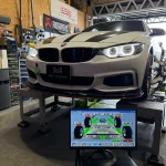 White BMW on a wheel-alignment dyno in a workshop, monitor showing alignment data in the foreground.