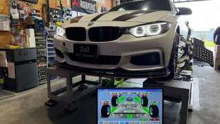 White BMW on a wheel-alignment dyno in a workshop, monitor showing alignment data in the foreground.