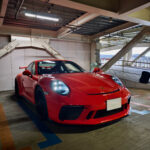 Red Porsche sports car parked in an indoor parking garage with exposed ceiling pipes above it.
