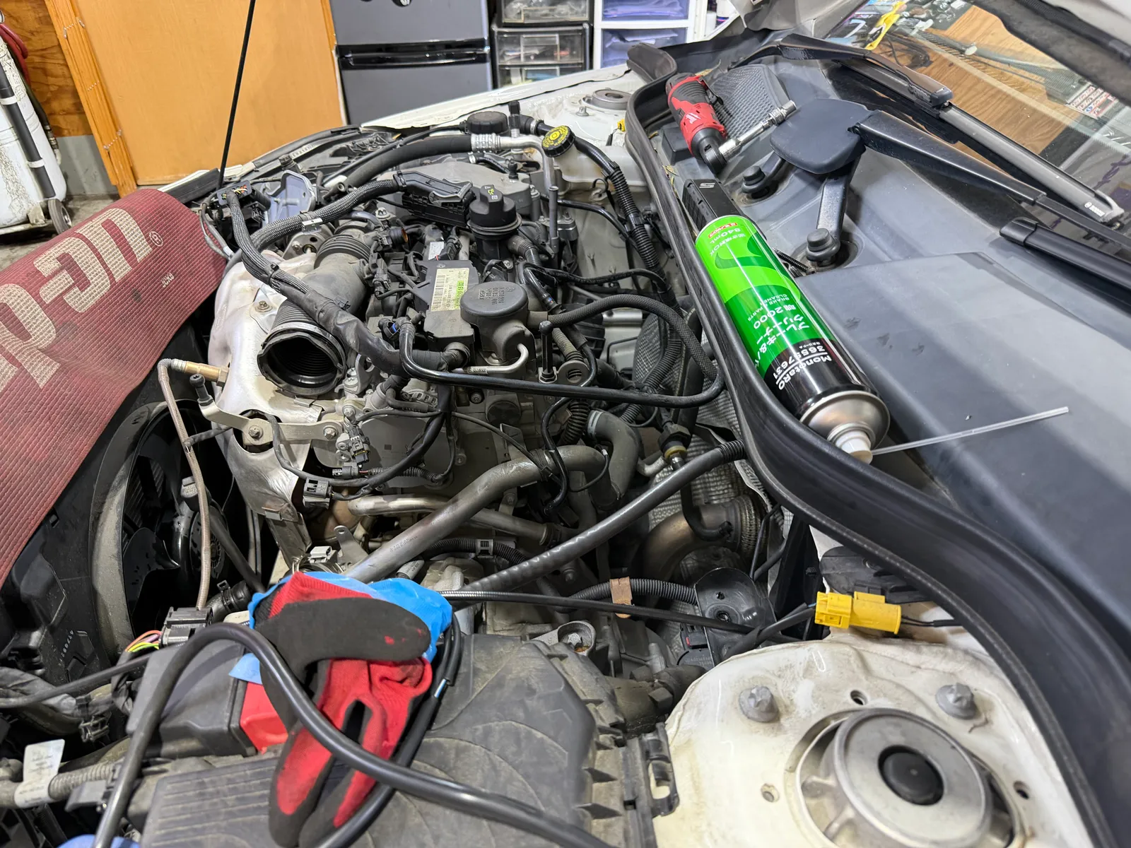Car engine bay with hood open and tools laid on the front, a green spray can resting on the fender and a red cordless drill nearby.