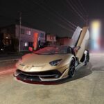 Gold Lamborghini with scissor doors open, parked on a city street at night, red interior glow visible.