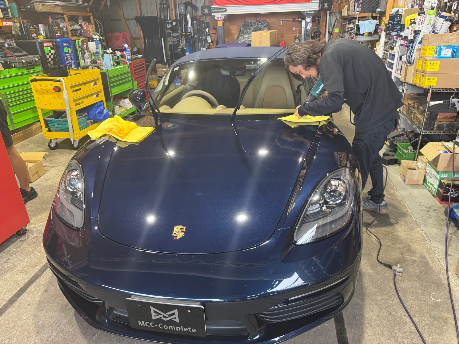 boxster718s-protection-film-2 – mbFAST Tuning Blue Porsche being polished in a cluttered workshop, with a person wiping the hood with a yellow microfiber cloths.