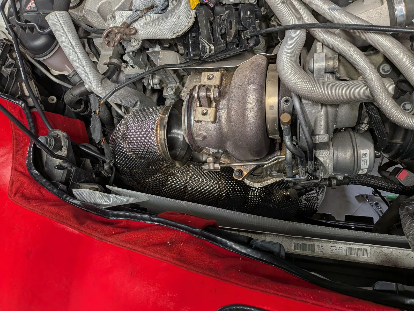 Close-up of a turbocharger in a car engine bay, with braided exhaust heat wrap and surrounding hoses and clamps visible.