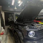 Black Mercedes-Benz G-Class SUV in a workshop with the hood raised, engine visible.