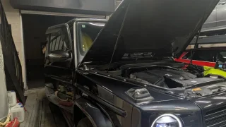 Black Mercedes-Benz G-Class SUV in a workshop with the hood raised, engine visible.
