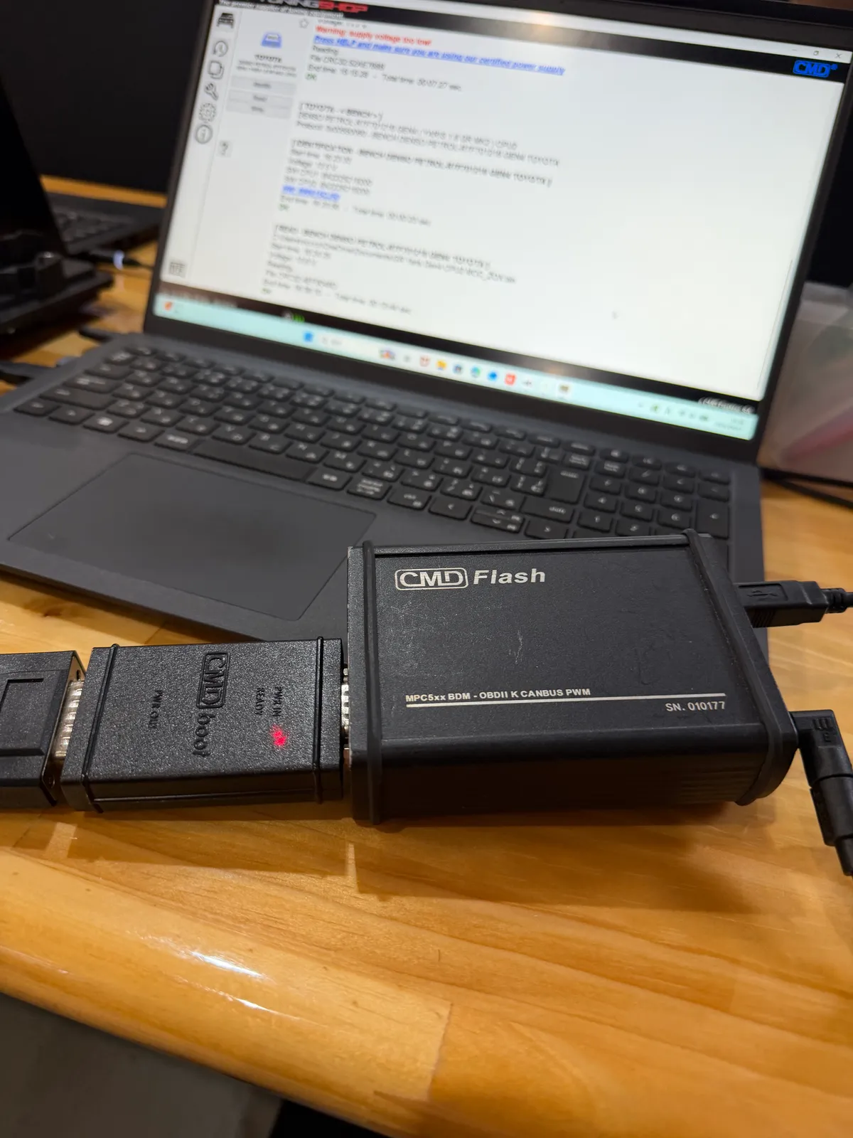 gryaris-ecu-tuning-2 – mbFAST Tuning CMD Flash hardware programmer connected to a laptop via USB, resting on a wooden desk with a cable plugged in. It’s a black rectangular device labeled CMD Flash with MPC5xx BDM-OBDII K CANBUS PWM text visible.
