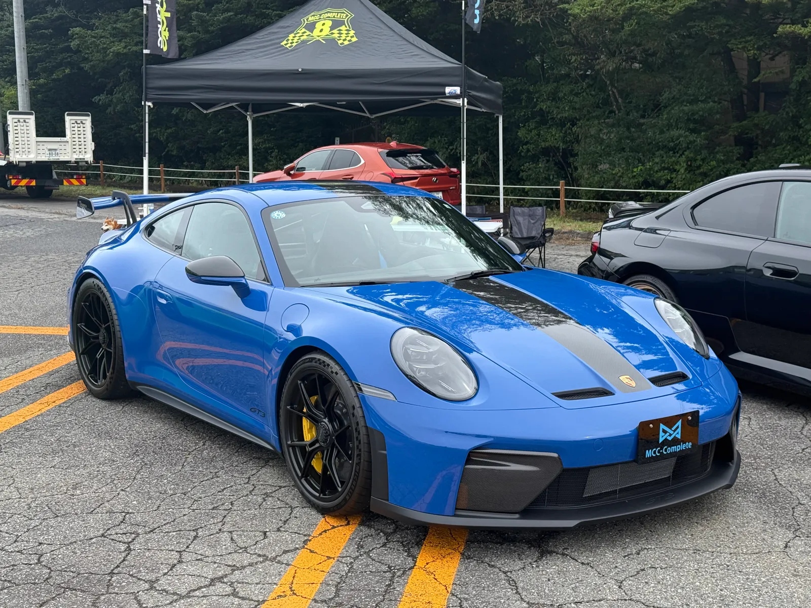 Blue Porsche 911 GT3 with a black center stripe and yellow brake calipers parked at a car show under a black tent.