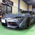 Front three-quarter view of a gray sports car inside a car workshop with tools and banners in the background, ready for service.