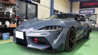 supra-a91-ecu-tuning-bubbling-1 – mbFAST Tuning Front three-quarter view of a gray sports car inside a car workshop with tools and banners in the background, ready for service.